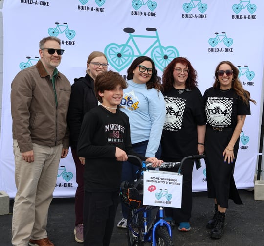 Student poses with new bike in front of ROE4 Student Services team members, who stand in front of a Build-a-bike backdrop